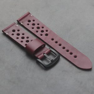 May include: A burgundy leather watch strap featuring a perforated design with multiple circular cutouts. The strap includes a matte black metal buckle and a matching leather keeper loop, with visible white stitching at the attachment points.