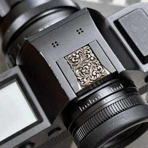 以下が含まれることがあります： 銀色の装飾的なフローラルデザインが施された黒いカメラのクローズアップ。 カメラにはレンズ、ダイヤル、ビューファインダーがあります。 デザインはカメラ本体の上部に配置されています。 写真撮影ツールです。