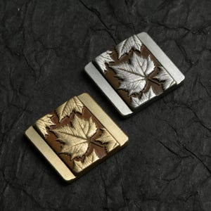 Könnte beinhalten: Zwei quadratische Metallschnallen mit Ahornblatt-Designs. Eine Schnalle hat eine silberne Oberfläche mit braunem Hintergrund und silbernen Blättern. Die andere hat eine goldene Oberfläche mit braunem Hintergrund und goldenen Blättern.