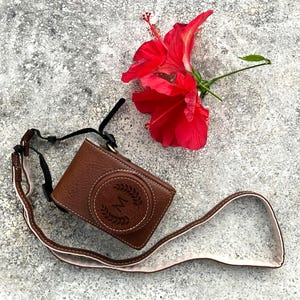 Może przedstawiać: Brązowe skórzane etui na aparat z pasującym paskiem, z okrągłym wzorem z literą "M" i wieńcem laurowym. W pobliżu spoczywa żywy czerwony kwiat hibiskusa.