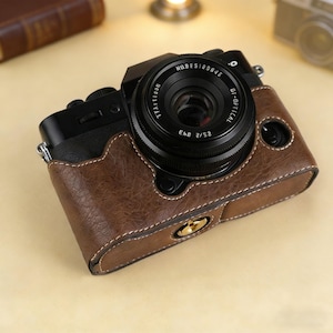 Puede incluir: Una c&aacute;mara digital negra con una lente, dentro de una funda de cuero marr&oacute;n. La c&aacute;mara tiene una fijaci&oacute;n de correa plateada y un cierre dorado. La lente tiene el texto "NORES189846" y "OI-OPTICAL".