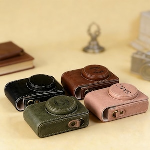 May include: Four leather camera cases in black, brown, green, and pink. Each case has a circular top with white stitching and a metal clasp. The brown and pink cases have initials engraved on the top. The cases are on a light yellow surface.