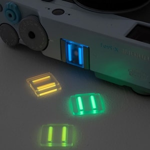 Può includere: Primo piano di una fotocamera grigio chiaro con accenti blu e verdi luminosi. Tre piccoli accessori rettangolari traslucidi con barre gialle e verdi luminose sono sparsi sotto.