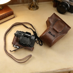 Könnte beinhalten: Eine schwarze Fujifilm X-T50 Kamera mit brauner Ledertasche und -riemen. Die Kamera ist teilweise in einer braunen Lederhülle eingeschlossen. Die passende Ledertasche ist offen, mit dem Namen "Mark" auf der Seite geprägt.