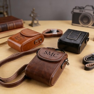 Może przedstawiać: Kolekcja skórzanych futerałów na aparat w różnych odcieniach brązu i czerni. Futerały mają białe przeszycia, zapięcia i paski. Jeden futerał jest wytłoczony inicjałami "SMC". Widoczny jest również pasek do aparatu i ilustracja aparatu w stylu vintage.