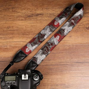 May include: A camera strap with a red, silver, and black butterfly pattern. The strap is made of a woven material and has black leather ends. The strap is attached to a black DSLR camera.