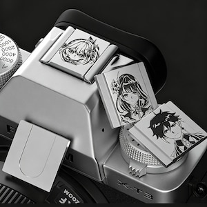 以下が含まれることがあります： アニメスタイルのキャラクターイラストが描かれた、3つの装飾的な金属プレートが付いたシルバーのカメラ。 カメラにはダイヤルとボタンがあり、プレートはホットシューやその他の場所に配置されています。