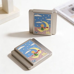 Peut inclure: Deux boîtes métalliques argentées avec une illustration colorée d'un jeune garçon sur un croissant de lune sous un ciel étoilé. Les boîtes sont rectangulaires avec un motif en relief sur le dessus.