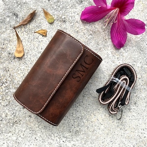 May include: A brown leather case with the initials "SMC" embossed on the front. The case is rectangular with a curved flap and white stitching. A black and tan strap with a silver clasp is next to the case. A pink flower and leaves are in the background.