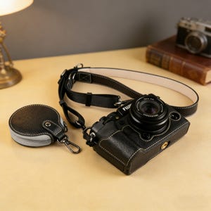 Könnte beinhalten: Eine schwarze Kamera mit Lederetui und -riemen, mit weißen Nähten. Ein passendes rundes Etui mit grauem Rand und silbernem Verschluss ist ebenfalls vorhanden. Das Kameraobjektiv ist schwarz, und der Riemen ist schwarz mit cremefarbener Unterseite.