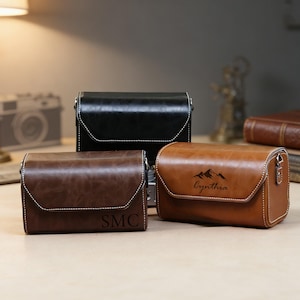 May include: Three leather camera cases in black, brown, and tan. The cases have white stitching and a flap closure. The brown case has the initials "SMC", and the tan case has a mountain graphic with the name "Cynthia".