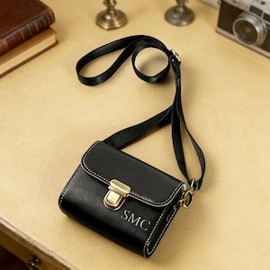 May include: Black crossbody bag with a flap closure and gold-tone hardware. The bag features white stitching and the initials "SMC" on the front. It has a black strap and is likely made of leather or a leather-like material.