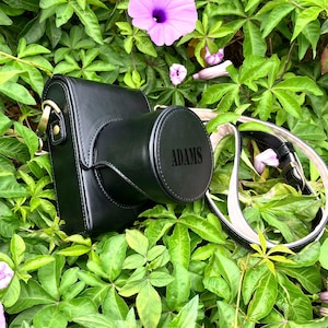 Può includere: Custodia per fotocamera in pelle nera con il nome "ADAMS" impresso sulla copertura dell'obiettivo. La custodia ha un cinturino nero abbinato con fibbia color oro. La custodia è circondata da foglie verdi e fiori viola.