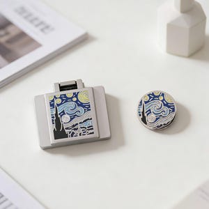 Peut inclure: Un support de téléphone rectangulaire argenté et une épingle ronde, tous deux ornés d'un motif inspiré de "La Nuit étoilée" de Van Gogh. L'œuvre présente des coups de pinceau bleus et jaunes tourbillonnants, une silhouette sombre de ville et un ciel étoilé.