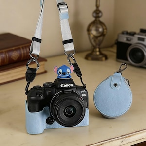 May include: A black Canon EOS R10 camera with a blue leather case and a matching strap. A blue Stitch figurine sits on top of the camera. A light blue round case with a zipper is also present.