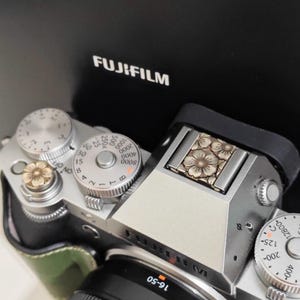 Könnte beinhalten: Silberne Fujifilm Kamera mit floralen Akzenten auf den Rädern und der Oberseite. Die Kamera hat ein schwarzes Objektiv und einen grünen Ledergriff. Das Fujifilm-Logo ist sichtbar. Das Objektiv liest 16-50.