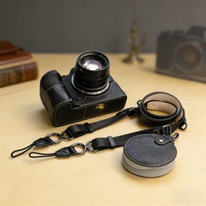 May include: A black leather camera with a lens attached, a matching strap, and a round case. The camera has white stitching and a textured surface. The strap and case are also black with silver hardware.