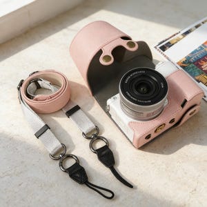 Può includere: Un set di custodia e cinturino per fotocamera in pelle rosa. La custodia è aperta, rivelando una fotocamera bianca e argento con un obiettivo nero. Il cinturino è rosa chiaro e grigio con dettagli neri e anelli metallici. Un accessorio elegante.