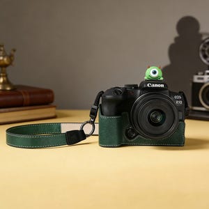 Puede incluir: Una c&aacute;mara Canon EOS R10 negra con una funda y correa de cuero verde. La lente de la c&aacute;mara est&aacute; centrada y una peque&ntilde;a figura de monstruo verde est&aacute; encima. La correa es verde oscuro con costuras blancas.