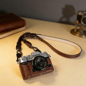Könnte beinhalten: Silberne Fujifilm-Kamera mit brauner Ledertasche und -riemen. Das Kameraobjektiv ist schwarz, und der Riemen ist braun mit weißen Nähten. Die Kamera liegt auf einer hellen Oberfläche.