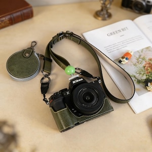 May include: A black Nikon Z series camera with a green leather case and strap. The camera has a lens attached and a green bead on the strap. A matching green leather pouch is attached to the strap. The camera is on a light-colored surface.