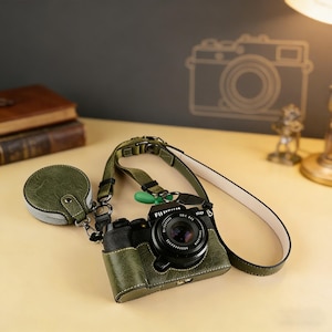 Könnte beinhalten: Eine grüne Kameratasche aus Leder mit passendem Objektivdeckel und Riemen. Die Kamera hat ein schwarzes Objektiv und ist mit "FUJIFILM" beschriftet. Die Tasche ist mit weißem Faden genäht. Der Riemen ist auf der Unterseite weiß.