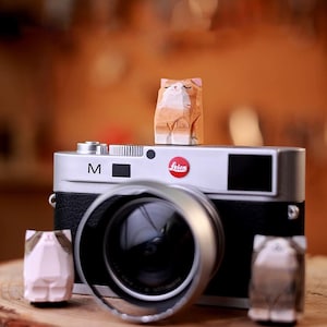 Könnte beinhalten: Eine silberne und schwarze Leica-Kamera mit Objektiv und drei kleinen Holzkatzenfiguren. Die Katzenfiguren sind in verschiedenen Farben, darunter Orange, Rosa und Grau. Die Kamera hat das Leica-Logo in Rot.