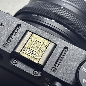May include: Close-up of a black camera with a lens and a gold-colored National Geographic logo. The logo is rectangular with the words "NATIONAL GEOGRAPHIC" printed below a square outline. The camera body is gray with various buttons and markings.