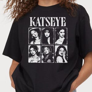 May include: A black short-sleeve t-shirt featuring the white text KATSEYE at the top. Below the text is a grid of six individual black and white portraits of different people, each framed in a rectangular box with a distressed, vintage-style print.