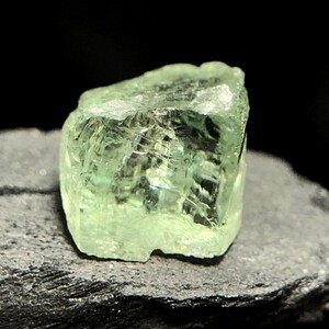 May include: A close-up of a rough-cut, light green gemstone. The gem has a crystalline structure with visible internal fractures and a slightly translucent appearance. The stone is resting on a dark, textured surface, with a black background.