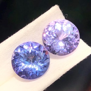 Può includere: Due gemme rotonde e sfaccettate con taglio brillante. Le gemme mostrano una gamma di tonalità blu e viola, con riflessi interni che creano un effetto scintillante. Riposano su una superficie bianca e strutturata su sfondo scuro.