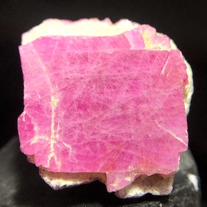 May include: A vibrant pink mineral specimen with a rectangular shape, showcasing a crystalline structure. The pink stone rests on a base of off-white material, creating a contrast. The mineral's surface displays fine lines and textures.