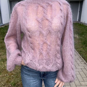 Könnte beinhalten: Ein helllila, durchsichtiger Strickpullover mit Zopfmuster auf der Vorderseite und den Ärmeln. Der Pullover hat lange, puffärmel und einen runden Ausschnitt. Die Person trägt eine blaue Jeans.