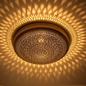 Può includere: Una plafoniera rotonda in metallo con un intricato motivo in filigrana tagliato al laser. La superficie metallica color oro presenta un denso motivo floreale ripetitivo che proietta un drammatico gioco di ombre sul soffitto.