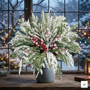 May include: A decorative arrangement of artificial evergreen branches covered in white flocking to resemble snow, interspersed with clusters of bright red berries. The foliage is contained within a dark gray ceramic vase with a textured pattern.