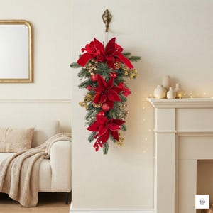 May include: A decorative teardrop-shaped wall hanging featuring deep red velvet poinsettia flowers, green pine branches, small red ornaments, and gold-toned accents. The arrangement is suspended from a metal hook on a light-coloured wall.
