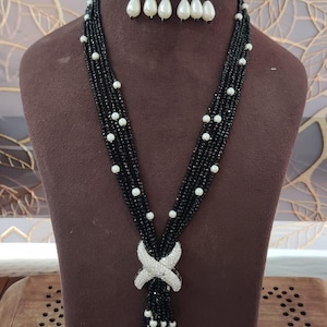 May include: A black beaded necklace and matching earrings set. The necklace features a silver-colored X-shaped pendant and white pearl accents. The earrings have a similar design with white pearl drops. The jewelry is displayed on a brown mannequin.