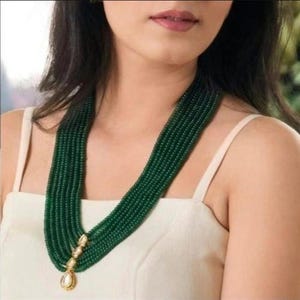 Puede incluir: Un collar de cuentas verde esmeralda de múltiples hilos con un colgante dorado. El collar está drapeado alrededor del cuello, mostrando el intrincado trabajo de cuentas y el diseño del colgante. El colgante tiene una perla en forma de lágrima.
