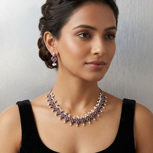 May include: A silver necklace and matching earrings set. The necklace and earrings feature a pattern of red gemstones and small white pearls. The jewellery is worn against a black top.