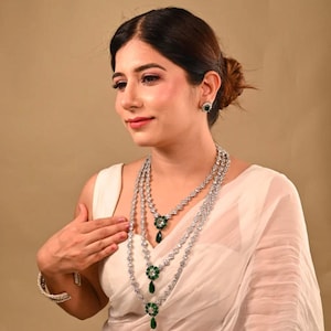 May include: A woman wearing a three-strand necklace, matching earrings, and a bracelet. The jewelry features clear stones and green accents. The woman is wearing a white top against a neutral background.