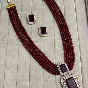 Puede incluir: Un conjunto de collar y pendientes de cuentas de color burdeos. El collar presenta múltiples hilos de pequeñas cuentas, con un gran colgante rectangular. Los pendientes también son rectangulares con un diseño similar. El conjunto está acentuado con metal dorado.