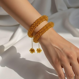 Puede incluir: Dos pulseras doradas con cuentas se llevan en una muñeca. Cada pulsera tiene una cadena con un pequeño dije de flor con cuentas. Las pulseras están hechas de pequeñas cuentas redondas. El fondo es una tela blanca drapeada.