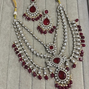 Peut inclure: Un ensemble de bijoux orné comprenant un collier, des boucles d'oreilles et un serre-tête. L'ensemble est argenté avec des pierres précieuses claires et rouges. Le collier a plusieurs couches avec un grand pendentif.