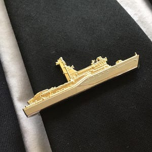 Può includere: Un fermacravatta dorato a forma di nave, appoggiato su una cravatta a righe bianche e nere. La clip presenta dettagli di una nave, tra cui una sovrastruttura e altri elementi.