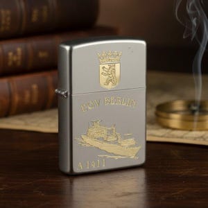 Może przedstawiać: Srebrna zapalniczka ze złotymi akcentami. Zapalniczka posiada herb, napis "EGV BERLIN", ilustrację statku i numer "A 1411". W tle widoczny jest dym.