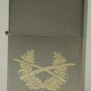 May include: A silver-colored metal lighter with a decorative emblem. The emblem features two crossed swords, a wreath, and oak leaves. The lighter has a rectangular shape with rounded edges.
