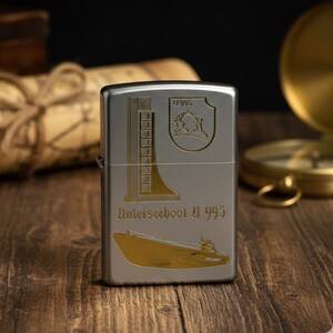 Könnte beinhalten: Ein silberfarbenes Feuerzeug mit goldfarbenen Grafiken eines U-Boots, eines Schildes und einer hohen Struktur. Der Text "U 995" und "Unterseeboot U 995" sind ebenfalls abgebildet. Das Feuerzeug liegt auf einer Holzoberfläche.