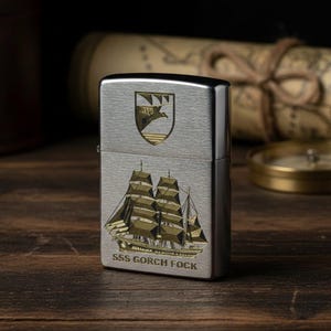 Può includere: Un accendino color argento con un'incisione dettagliata di una nave a vela e la scritta "SSS GORCH FOCK". L'accendino presenta anche un disegno a stemma. Sullo sfondo sono visibili una mappa e una bussola.