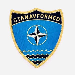 Könnte beinhalten: Blau-goldener, gestickter Aufnäher mit dem Text "STANAVFORMED". Er zeigt eine weiße und schwarze Windrose in einem Kreis und Wellen am unteren Rand. Der Aufnäher hat einen goldenen Rand.