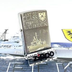 May include: A silver Zippo lighter with the text "LAST OF CLASS Fregatte LÜBECK" and a ship engraving. The lighter is displayed on a clear stand with the Zippo logo. The background features a ship illustration.
